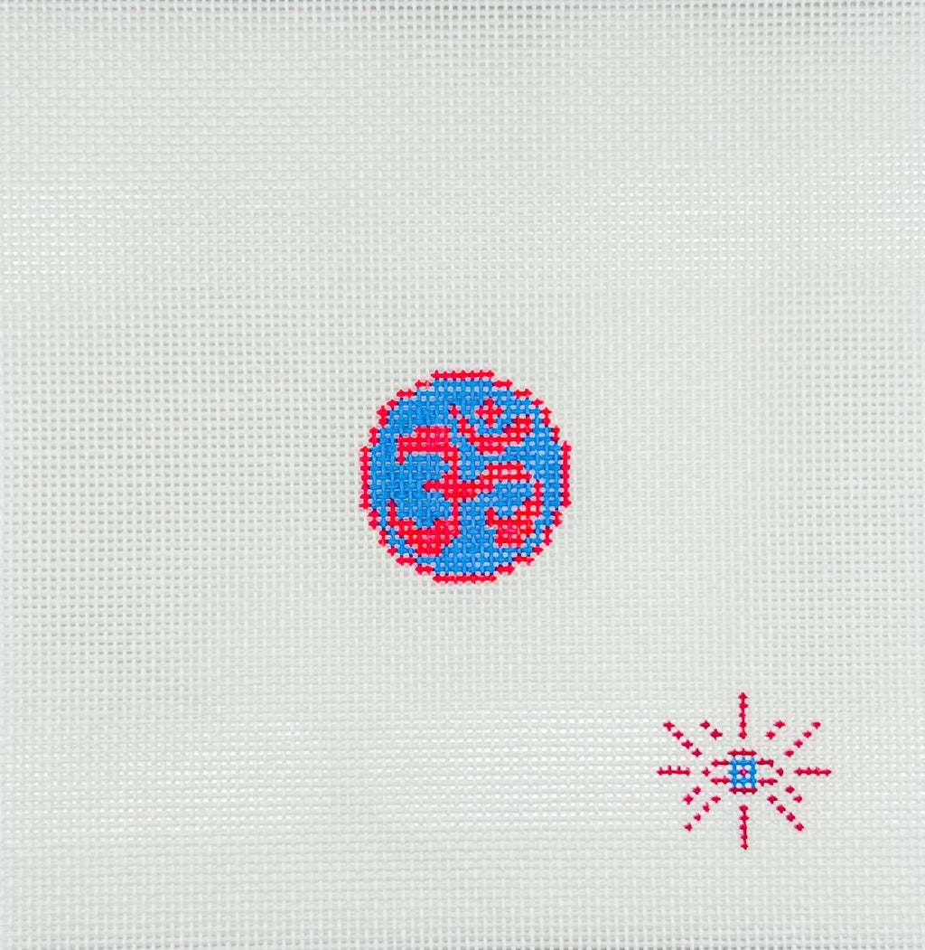 A close-up of white Aida cloth canvas featuring a small embroidered Om symbol in blue and red, with a smaller red and blue starburst motif in the lower right corner.